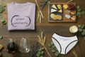Wine & Cheese Experience Gift Set flat lay with purple tee, stemless wine glasses, engraved acacia charcuterie board and briefs